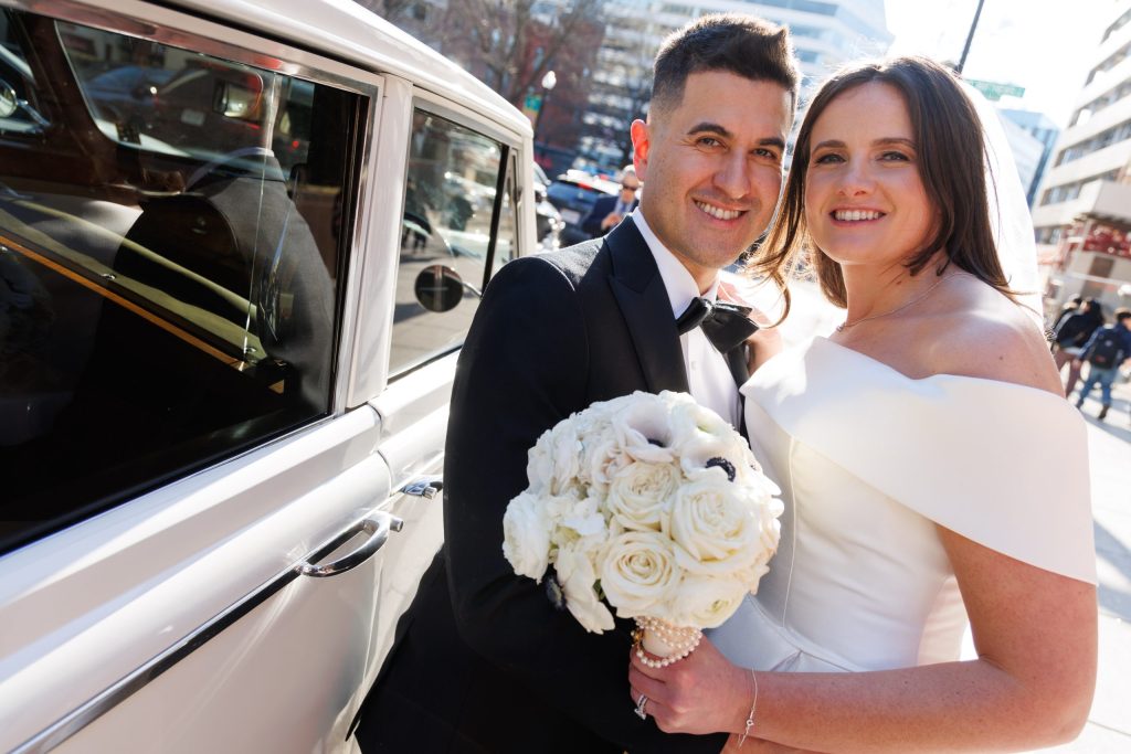 Bride and groom smiling together beside a vintage car on a city street