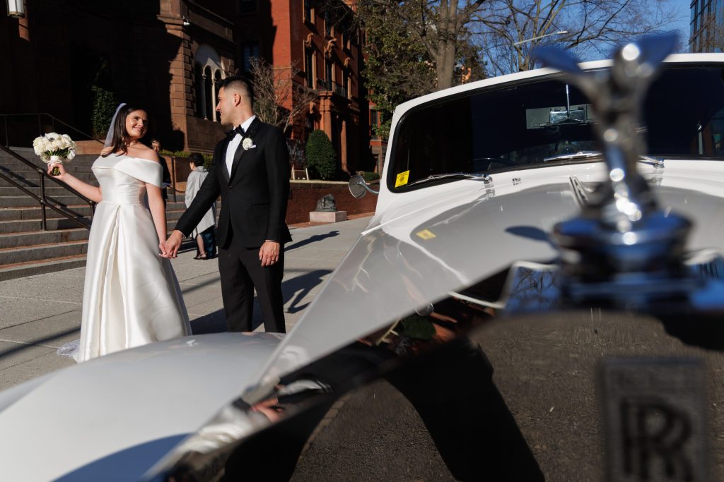 Bride and groom holding hands as they walk toward a vintage car outside a city church
