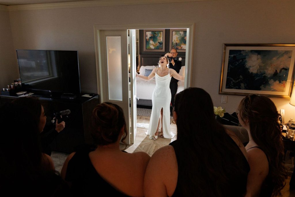 Bride laughing joyfully as she shows off her wedding dress to bridesmaids while getting ready